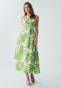 Maxi haljina od bijelog materijala s velikim zelenim uzorcima lišća. Užitak u struku, bez rukava, lepršava suknja, uparena s bijelim sandalama.