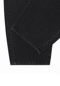 Deux jambes de pantalon en denim noir qui se chevauchent montrant des coutures détaillées sur les ourlets sur un fond blanc.