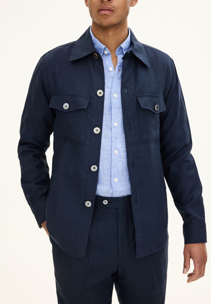 Navy linen button-up jacket with two chest pockets and silver buttons, worn over a light blue collared shirt, creating a layered look.