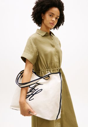 Mujer con cabello rizado que lleva un vestido de botones color oliva y sostiene una gran bolsa de lona blanca con letras negras y detalles en negro.