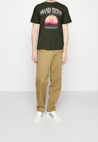 Camiseta de algodón verde oscuro con gráfico de "Gran Parque Nacional Teton" en rosa y morado. Combinada con pantalones beige claros y zapatillas blancas.