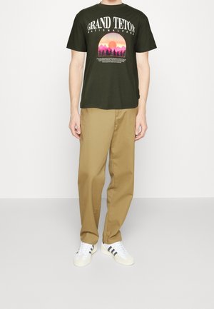 Mørkegrøn bomulds t-shirt med grafik af "Grand Teton National Park" i pink og lilla. Sammen med lyse beige bukser og hvide sneakers.