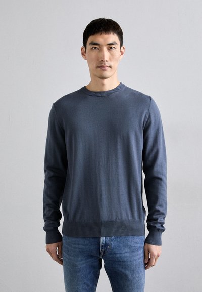 Maglione girocollo blu scuro con polsini e orlo a costine, realizzato in tessuto morbido, abbinato a jeans attillati blu chiaro.