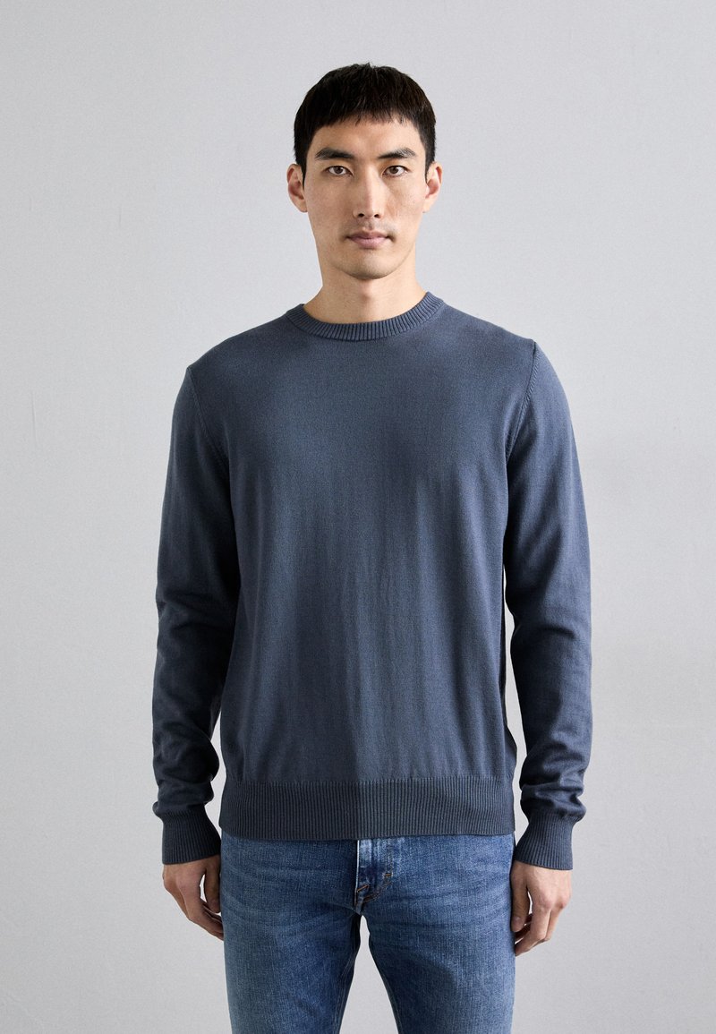 Pull bleu marine à col rond avec des poignets et un ourlet côtelés, en tissu lisse, associé à un jean ajusté bleu clair.