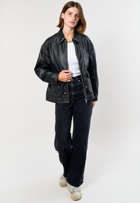 Giacca in pelle nera dal fit rilassato, con colletto a punta, chiusura con zip e cuciture visibili. Abbinata a jeans neri a vita alta e sneakers.