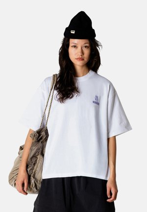 Młoda kobieta z długimi ciemnymi włosami, nosząca czarną czapkę beanie, oversize biały T-shirt z logo, czarne spodnie oraz beżową torebkę na ramię.
