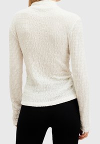 Valkoinen pitkähihaiseksi paita, jossa on teksturoitu aallokkokuosi. Pehmeä kangas, istuva muotoilu, pyöristetty helma ja korkea kaula-aukko. Yhdistetty mustien housujen kanssa.