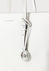 Mala tote de couro branca com superfície texturizada, alças duplas e um acréscimo metálico circular prateado com o nome da marca.