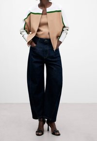 Giacca bomber color block beige e bianca con dettagli verdi, abbinata a pantaloni in denim scuri a gamba larga e sandali neri con tacco.