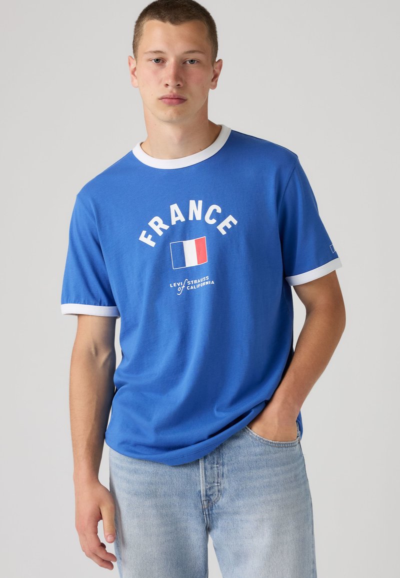 Giovane uomo in piedi con la mano in tasca, indossa una maglietta blu con bordino bianco, scritta "FRANCE", bandiera francese e jeans azzurri chiari.