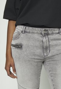 Jeans de ganga cinzentos com um corte slim, apresentando um bolso com fecho de correr na coxa esquerda e um leve efeito de desvanecimento. Cintura alta com um fecho de botão clássico.