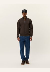 Καφέ μπουφάν με φερμουάρ και ψηλό γιακά, συνδυασμένο με ανοιχτόχρωμα τζιν και καφέ παπούτσια. Μαύρο καπέλο με λογότυπο στο κεφάλι.