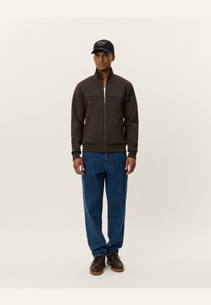 Brown zip-up jacket with a high collar, paired with blue loose-fitting jeans and brown shoes. Black cap with logo on head.