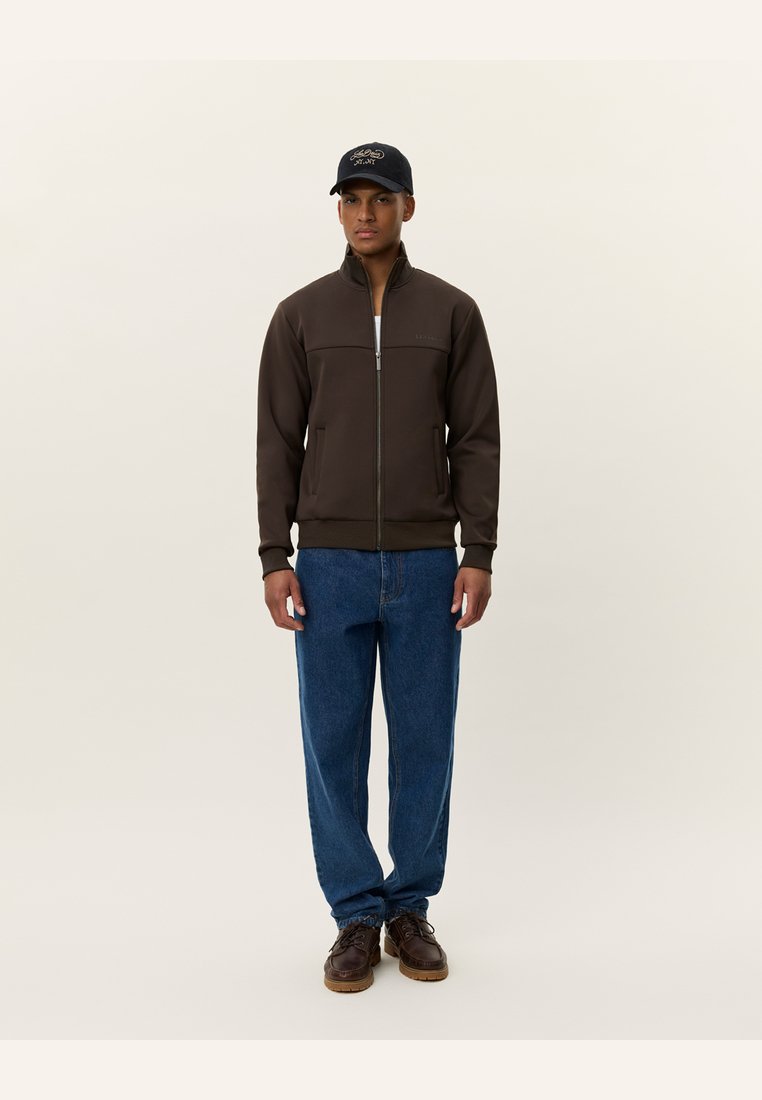 Καφέ μπουφάν με φερμουάρ και ψηλό γιακά, συνδυασμένο με ανοιχτόχρωμα τζιν και καφέ παπούτσια. Μαύρο καπέλο με λογότυπο στο κεφάλι.