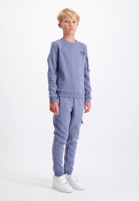 Jongen met blond haar, gekleed in een blauwe sweatshirt en bijpassende trainingsbroek, staand met armen langs zijn lichaam en witte sneakers op een witte achtergrond.