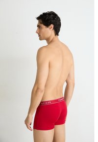 Piros pamut boxer alsónemű fekete és fehér "TOMMY HILFIGER" logós derékszorítóval, hátulról nézve, sima háttér előtt.
