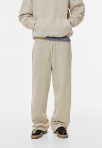 Beige kapucnis pulóver elülső zsebből, kék és fehér csíkos textil fölé rétegezve. Beige sportnadrág, húzózsinórral a derékban és szükülő lábakkal.