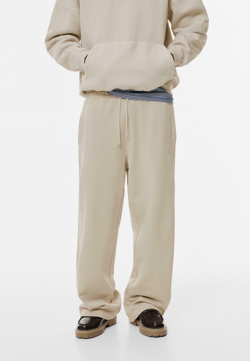 Beige kapucnis pulóver elülső zsebből, kék és fehér csíkos textil fölé rétegezve. Beige sportnadrág, húzózsinórral a derékban és szükülő lábakkal.