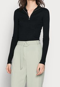 Haut noir côtel é à manches longues avec col zippé, associé à un pantalon taille haute vert clair avec ceinture en tissu et détails de passants.