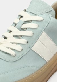 Baskets en daim bleu clair avec lacets blancs, bande latérale en cuir blanc et semelle en caoutchouc marron, vue en gros plan.