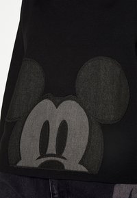 Černá mikina s velkou vyšívanou tváří Mickey Mouse v odstínech šedé, s aplikovanými kulatými ušima a prošitými detaily.