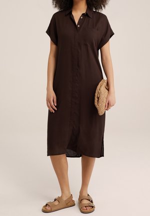 Frau trägt ein lockeres, dunkelbraunes Midi-Kleid mit Knöpfen und kurzen Ärmeln, hält eine gewebte Clutch und trägt hellbraune Sandalen mit Schnallen.