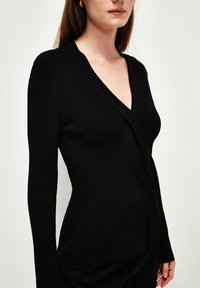 Zwarte geribbelde cardigan met een diepe V-hals, lange mouwen en een wikkelontwerp. De stof heeft een zachte, textuurachtige uitstraling.