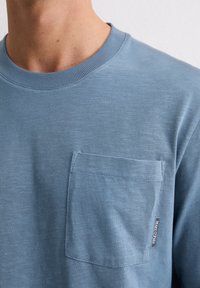 Jasnoniebieska koszulka z krótkim rękawem i okrągłym dekoltem, wykonana z teksturowanej tkaniny. Posiada kieszeń na piersi oraz małą metkę z logo marki na dolnej krawędzi kieszeni.