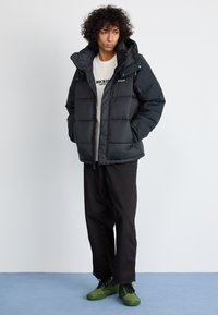Sort pufferjakke med hætte og quiltet design, båret over en grå skjorte og parret med sorte bukser og grønne sneakers.