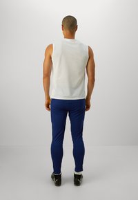 Αμάνικη λευκή αθλητική μπλούζα συνδυασμένη με μπλε εφαρμοστά παντελόνια. Η πλάτη αποκαλύπτει υφή από ύφασμα, ενώ τα παντελόνια έχουν φερμουάρ στους αστραγάλους.