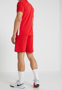 Camiseta y pantalones cortos deportivos rojos, hechos de una tela transpirable, con acentos laterales blancos. Combinados con zapatillas deportivas grises y rojas y calcetines blancos.