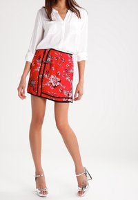 Femme portant une chemise blanche à boutons, une jupe courte à fleurs rouges avec des bordures noires, et des sandales à talons aiguilles argentées, se tenant devant un fond neutre.