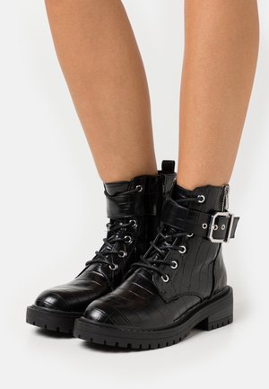 Černé kotníkové boty z kůže s krokodýlím vzorem, se šněrováním vpředu, bočním zipem, robustní podrážkou a kovovou přezkou na kotníku.