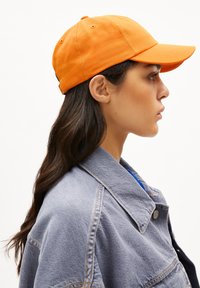 Orangene Baseballmütze mit glatter Textur, gebogenem Schirm und gestickten Ösen, getragen mit einer hellblauen Jeansjacke.