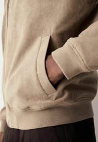 Beige ruskindsjakke med lynlåslukning, som har en kængurulomme og ribbede manchetter. Blød tekstur og en afslappet pasform.