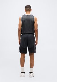 Άνδρας που στέκεται με την πλάτη γυρισμένη, φοράει μαύρο αμάνικο αθλητικό μπλουζάκι από δίχτυ, μαύρο αθλητικό σορτς, άσπρες κάλτσες και λευκά αθλητικά παπούτσια.