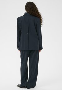 Sieviete ar gariem cirtainiem matiem, ģērbusies tumšā pārāk lielā uzvalkjakā un atbilstošās platajās biksēs, stāv ar muguru pret kameru uz baltas fona.