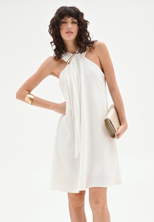 Femme aux cheveux bouclés et foncés portant une robe blanche sans manches avec une boucle dorée, un bracelet manchette doré, et tenant une pochette dorée.