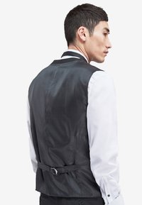 Schwarze Weste mit strukturiertem Finish, taillierte Passform, verstellbarer Rückenriemen, kombiniert mit einem weißen Hemd. Keine sichtbaren Muster oder Beschläge.