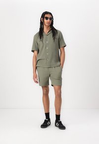 Olijfgroen kortomarmig overhemd met knopen aan de voorkant en bijpassende shorts. Wafelstructuur, gecombineerd met zwarte sneakers en een zonnebril.