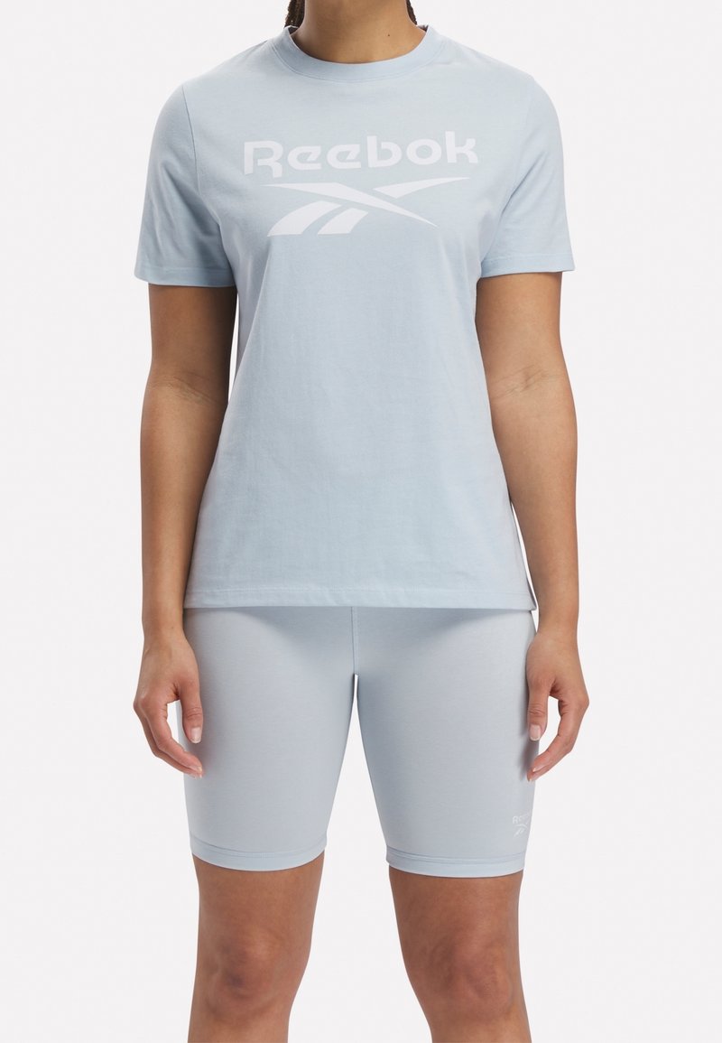 Personne portant un t-shirt Reebok bleu clair et un short ajusté assorti, debout devant un fond uni.
