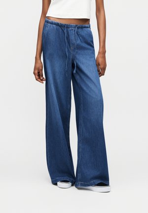 Γυναίκα που φορά χαλαρό μπλε τζιν παντελόνι με ελαστική μέση και κορδόνι, συνδυασμένο με μαύρα και λευκά αθλητικά παπούτσια.