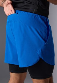 Zili sporta šorti zili ar vieglu, elpojošu materiālu. Iekļauj sānu kabatu un slāņotu dizainu virs melniem kompresijas šortiem.