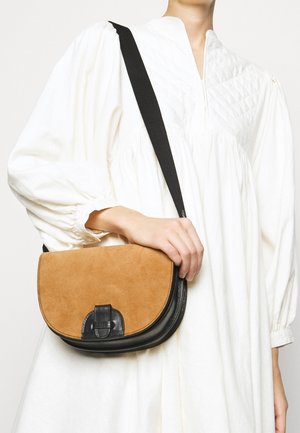 Personne en robe blanche à manches longues tenant un sac bandoulière beige et noir avec une large sangle noire.