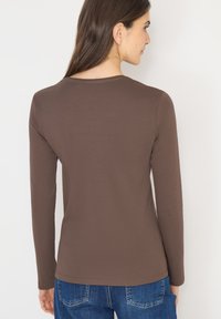Hnědý přiléhavý top s dlouhými rukávy a kulatým výstřihem, vyrobený z hladkého materiálu. Nošený s modrými džínami, představující jednoduchý design.
