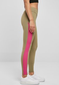 Olivengrønne leggingser med en lys pink sidepanel, som har en tettsittende design og elastisk midjebånd, sammen med hvite joggesko.