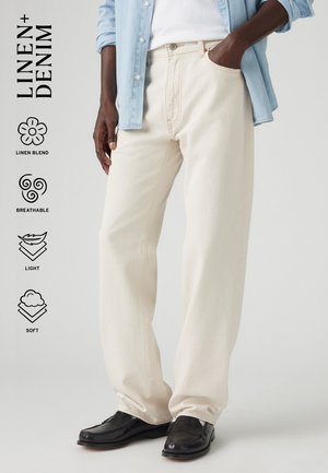 Personne portant un pantalon large en lin mélangé beige clair, un haut blanc, une chemise en denim bleu clair et des mocassins noirs, avec les qualités du tissu listées sur le côté.