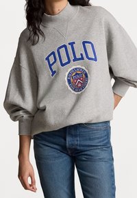 Felpa grigia con colletto alto, testo blu "POLO" e logo circolare con "Ralph Lauren" al centro. Design oversize.