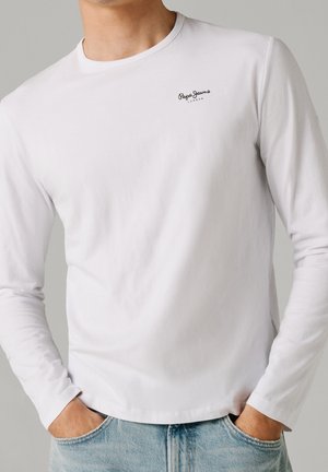 Mężczyzna w białej, długorękawowej koszuli Pepe Jeans London, z rękami w kieszeniach jasnoniebieskich jeansów, na jednolitym szarym tle.