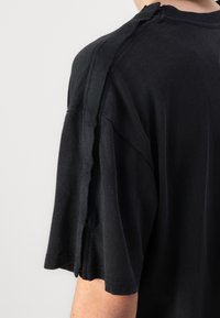 Gros plan d'une chemise noire à manches courtes avec un détail de fermeture éclair verticale courant le long de la couture de l'épaule et de la manche.